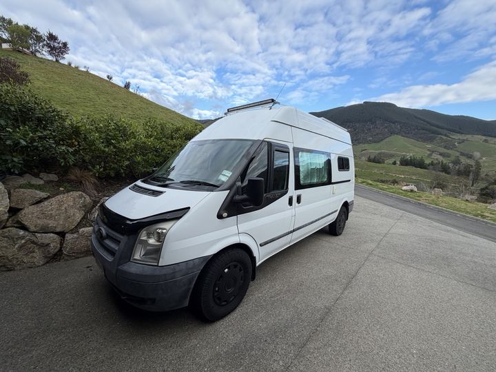 Front three-quarter view of the camper with green hills and cloud behind