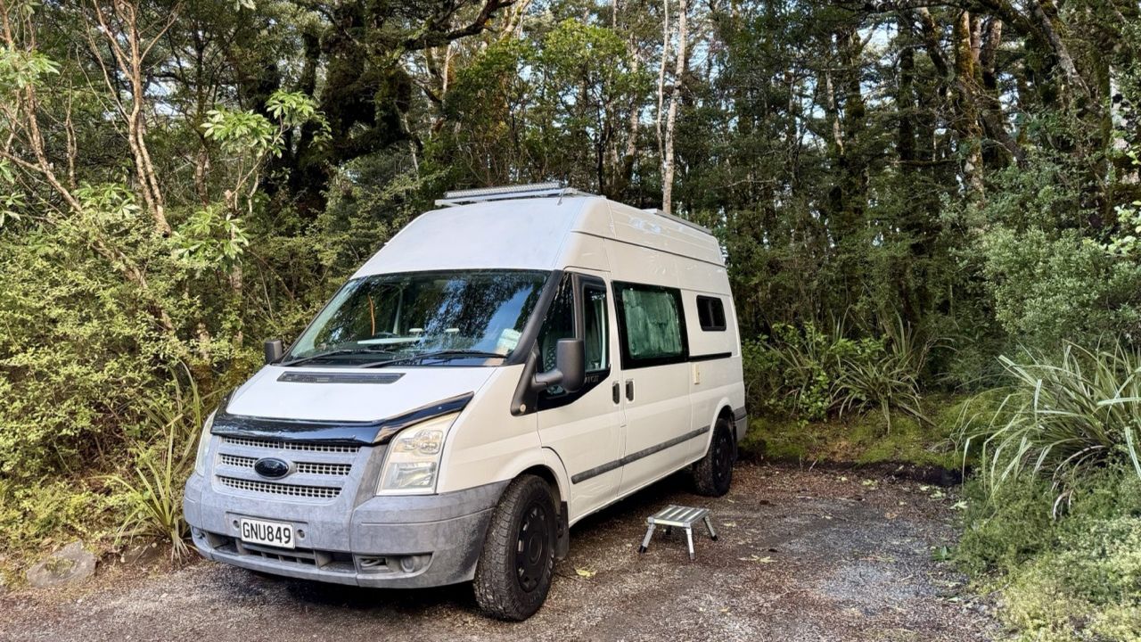 The white high-roof Transit camper parked on gravel in beech forest, step out by the sliding door